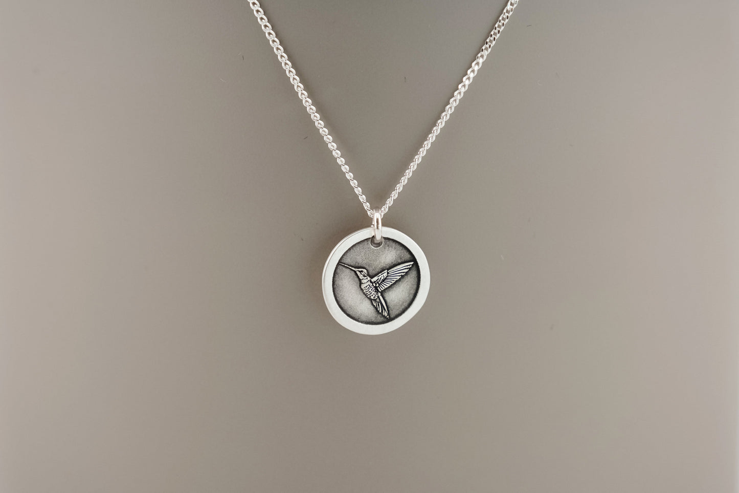 Silver necklace with a round pendant engraved with a hummingbird resting on a beige background