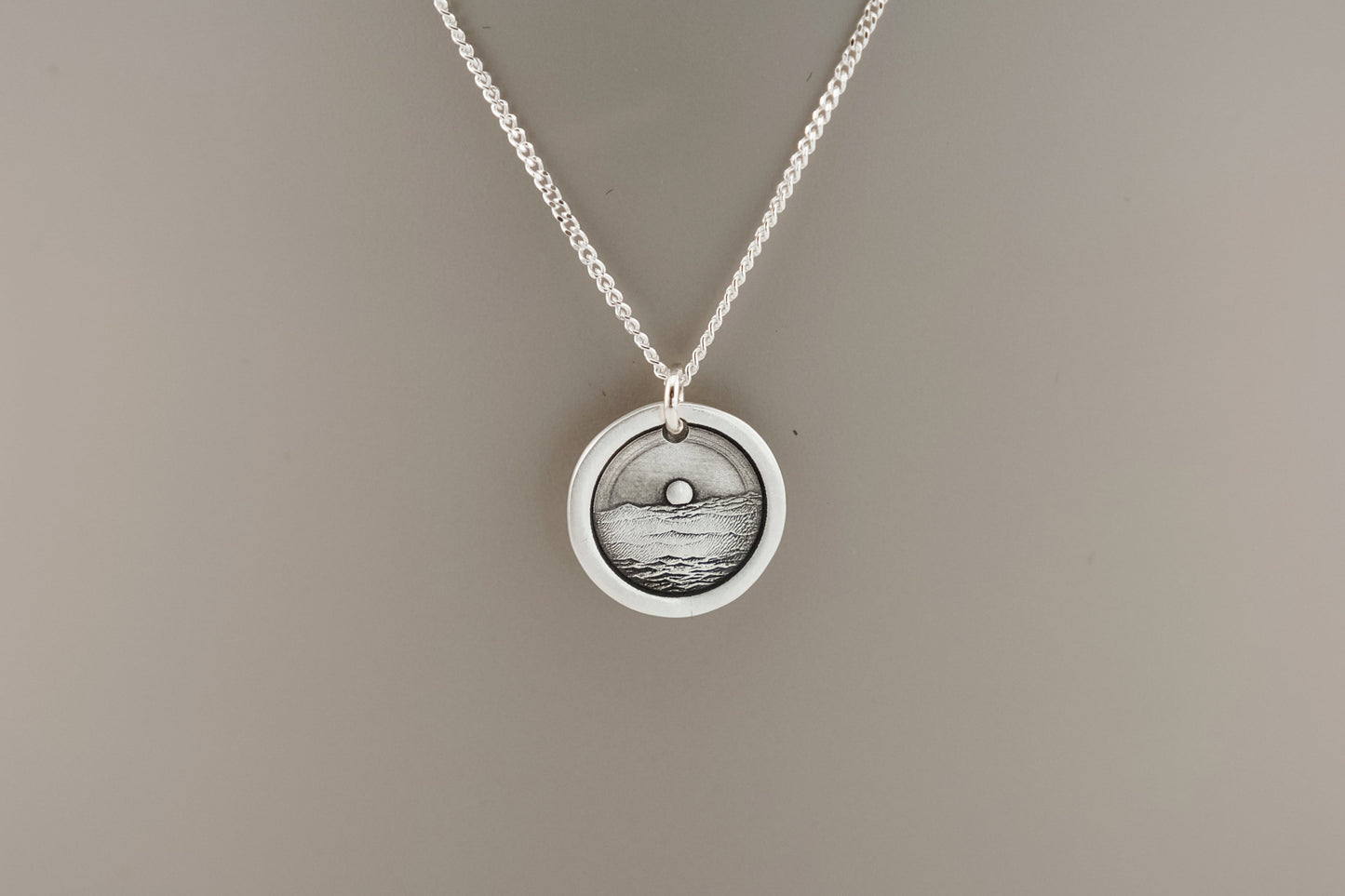 Silver necklace with a round pendant engraved with an ocean sunset on a plain background