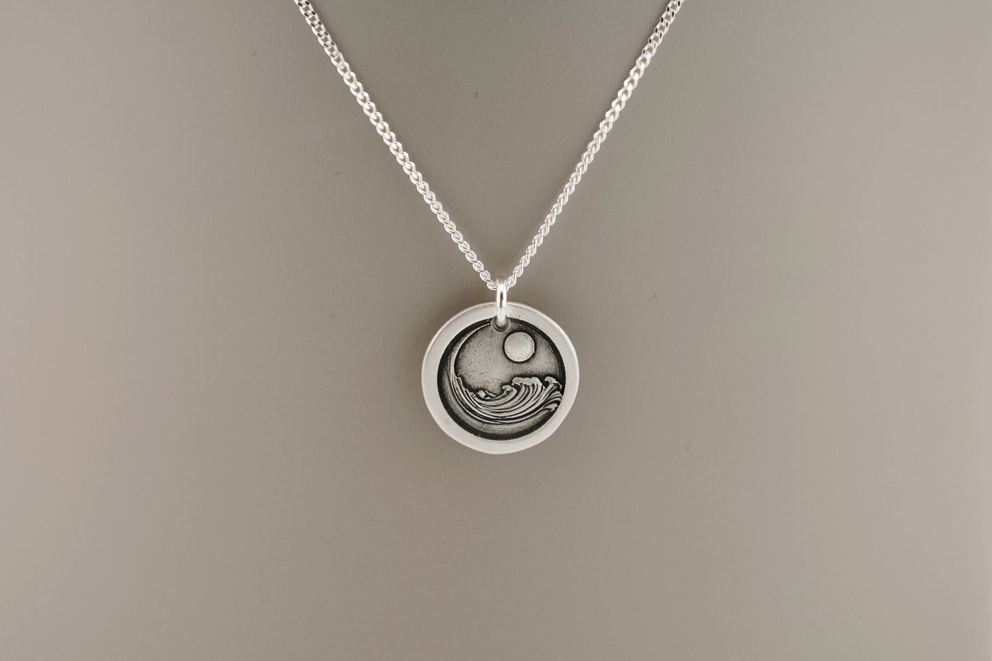 Silver necklace with a circular pendant engraved with an ocean wave resting on a plain background