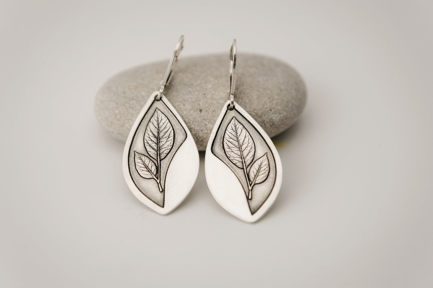 Silver teardrop shaped earrings featuring an engraved leaf design, resting on a stone surface.