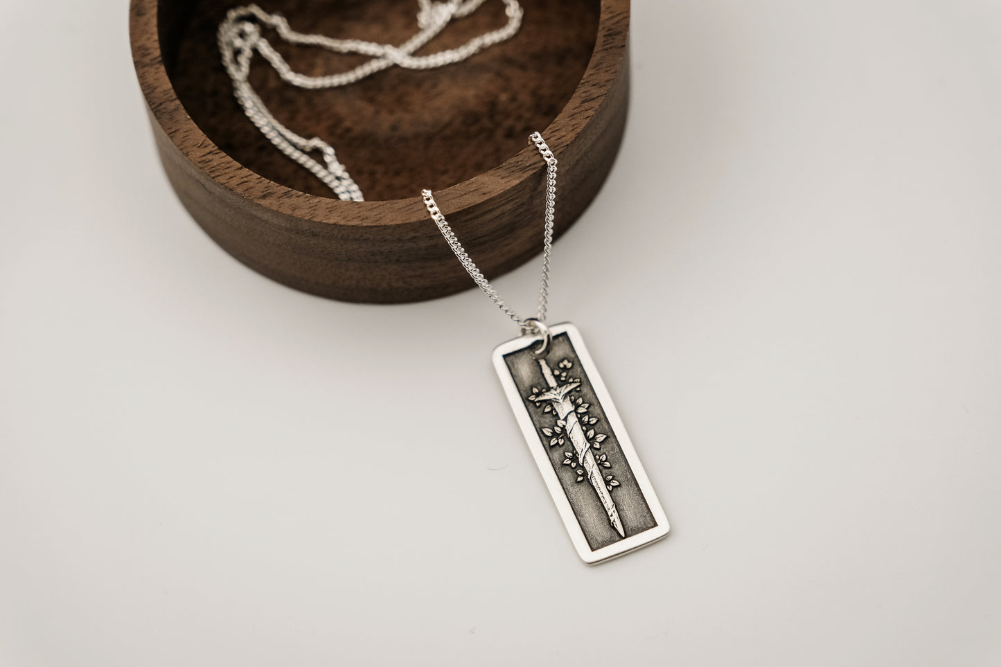 Silver necklace with a rectangular pendant featuring an engraved vine-wrapped sword resting on a light gray background