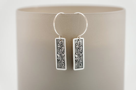 Silver earrings with engraved intricate botanical designs on a beige background
