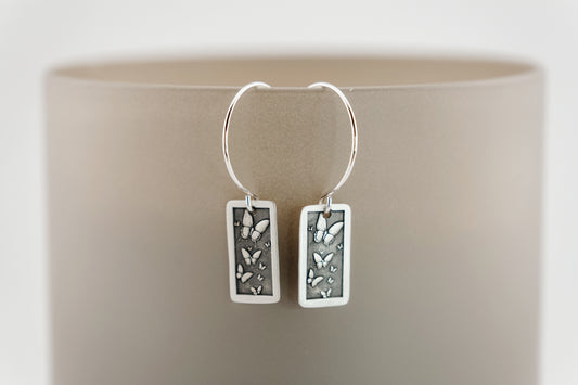 Silver hoop earrings with rectangular charms featuring engraved butterflies, hanging from on a beige background
