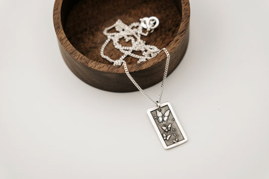 Silver necklace with a rectangular pendant engraved with butterflies laying on a wooden dish