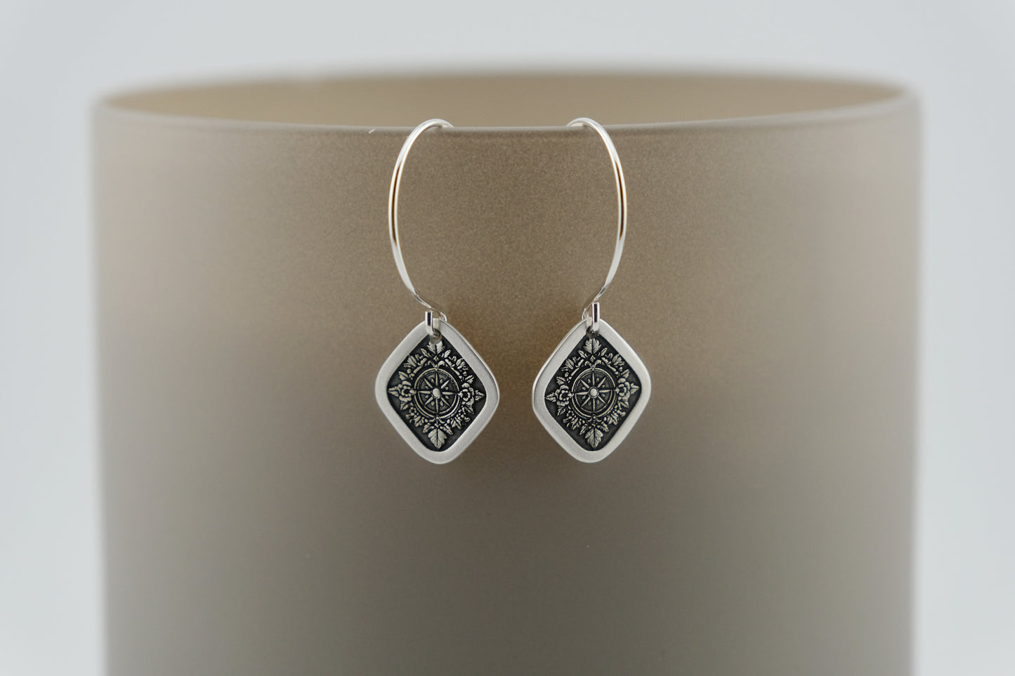 Silver hoop earrings with a diamond shaped charm featuring and engraving of a compass on a neutral background.