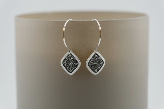 Silver hoop earrings with a diamond shaped charm featuring and engraving of a compass on a neutral background.