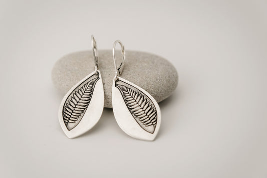 Silver leaf-shaped earrings featuring an engraved fern design on a gray stone surface