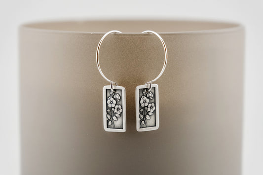 Silver hoop earrings with rectangular charm featuring flowers and a sword on a neutral background.
