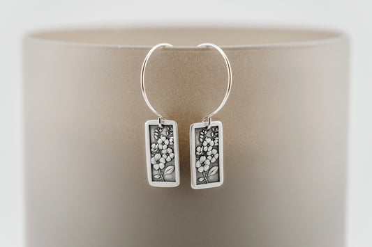 Silver hoop earrings with rectangular charm featuring engraved flowers on a neutral background.