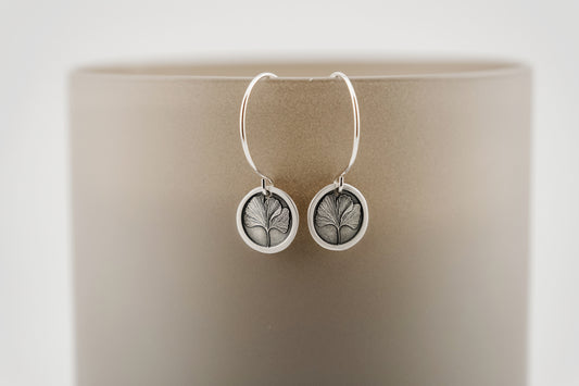 Silver earrings with ginkgo leaf design set against a beige surface