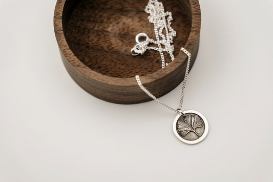 Silver necklace with an engraved ginkgo leaf circular pendant on a white surface with a wooden dish.