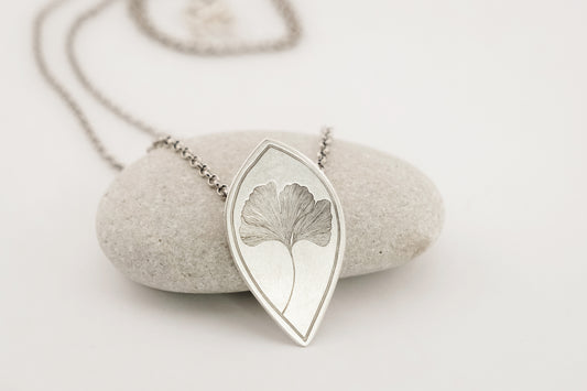 An engraved ginkgo leaf necklace made of sterling silver, displayed on a white stone. The pendant is oxidized and has a satin finish, hanging from an 18-inch sterling silver oxidized rolo chain with a hidden bail.