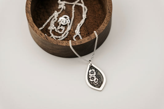 Silver necklace with a teardrop pendant engraved with a floral design in a wooden jewelry box on a light background