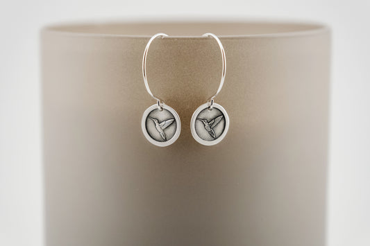 Silver earrings with circular design containing an engraved hummingbird, set on a beige background