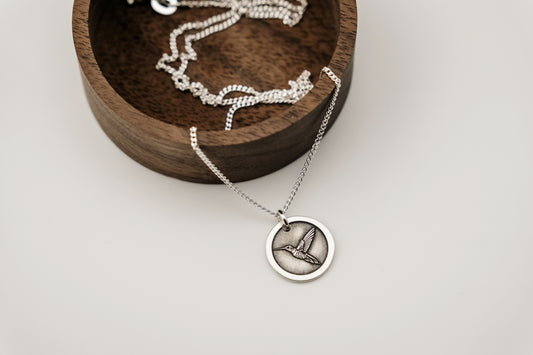 Silver necklace with a round pendant engraved with a hummingbird resting on a wooden tray