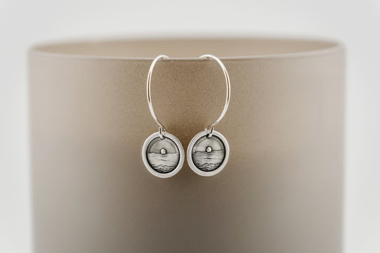 Silver earrings with circular design engraved with a scene of the sun dropping below the ocean, set against a beige background