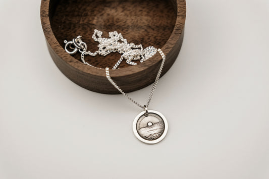 Silver necklace with a round pendant engraved with an ocean sunset resting on a wooden box