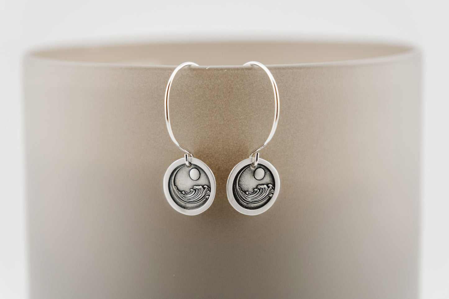 Silver earrings with circular design containing an engraving of a large ocean wave circling the moon, set on a neutral background 