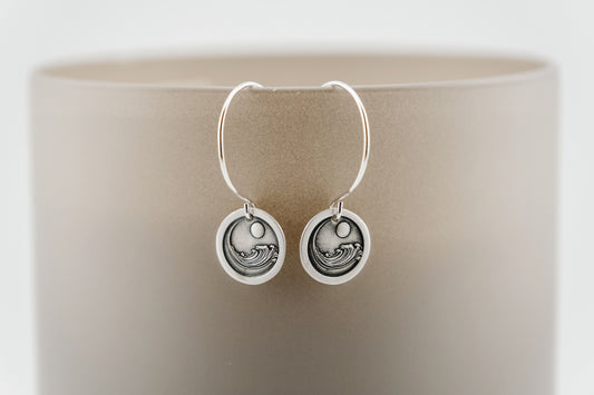 Silver earrings with circular design containing an engraving of a large ocean wave circling the moon, set on a neutral background 