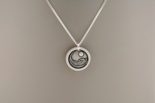 Silver necklace with a circular pendant engraved with an ocean wave resting on a plain background