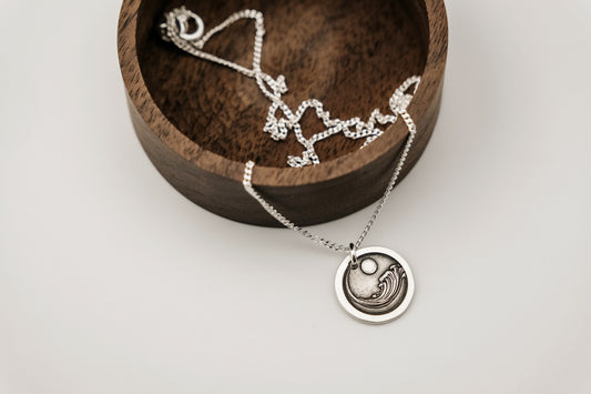 Silver necklace with a circular pendant featuring an engraving of an ocean wave resting on a wooden display box against a white background