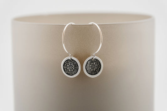 Silver hoop earrings with a round charm featuring an engraving of a compass on a neutral background.