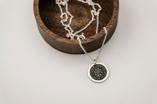 Silver necklace with a round compass pendant on a wooden dish against a white background