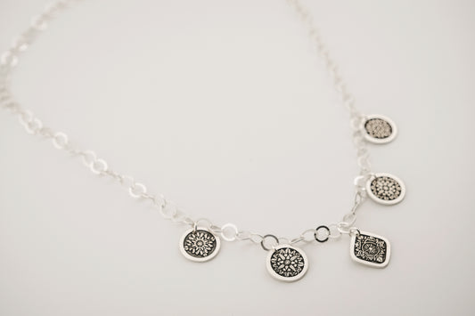 A silver charm necklace featuring circular and diamond shaped charms engraved with compass designs, resting on a white background