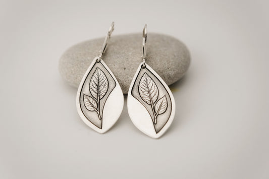 Silver teardrop shaped earrings featuring an engraved leaf design, resting on a stone surface.