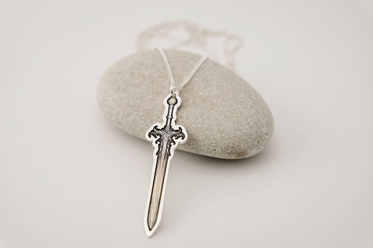 Silver sword-shaped pendant on a chain, placed on a light stone surface.