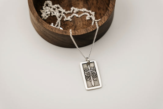 Silver necklace with a rectangular pendant featuring a sword and flowers on a light gray background