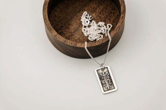 Silver necklace with a pendant engraved with a sword and flower design resting on a wooden stand against a white background