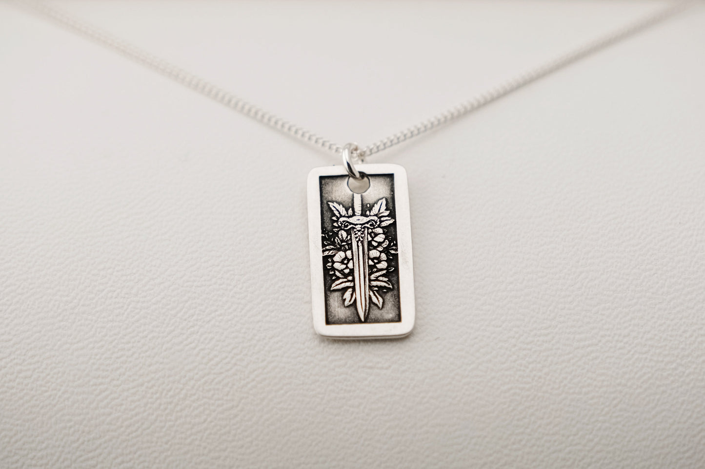 Silver necklace with a rectangular pendant featuring a sword and flowers on a light gray background