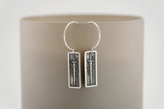 Silver hoop earrings with rectangular charms featuring a botanical vine wrapped around a sword on a neutral background.