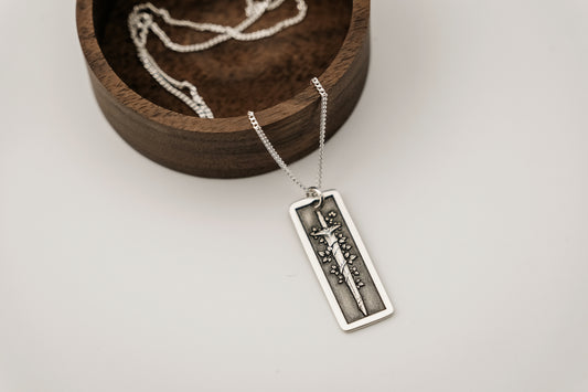 Silver necklace with a rectangular pendant featuring an engraved vine-wrapped sword resting on a light gray background