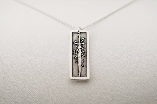 Silver necklace with a rectangular pendant featuring an engraved vine-wrapped sword resting on a light gray background