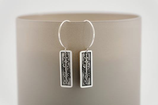 Silver hoop earrings with rectangular charms featuring a botanical vine wrapped around a sword on a neutral background.