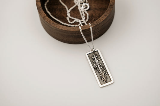 Silver necklace with a rectangular pendant engraved with a vine wrapped sword resting on a wooden dish