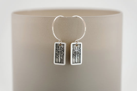 Silver hoop earrings with rectangular charm featuring flowers and a sword on a neutral background.