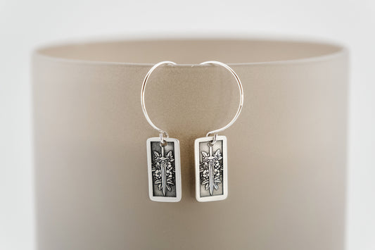 Silver hoop earrings with rectangular charm featuring flowers and a sword on a neutral background.