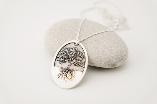 Silver necklace with an oval pendant featuring and engraved tree of life design resting on a white background