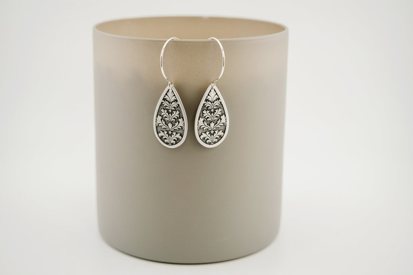 Teardrop-shaped silver earrings with gemstones on a beige cylindrical container against a light gray background