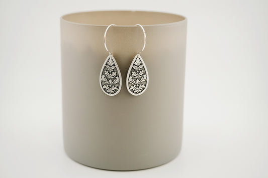 Teardrop-shaped silver earrings with gemstones on a beige cylindrical container against a light gray background