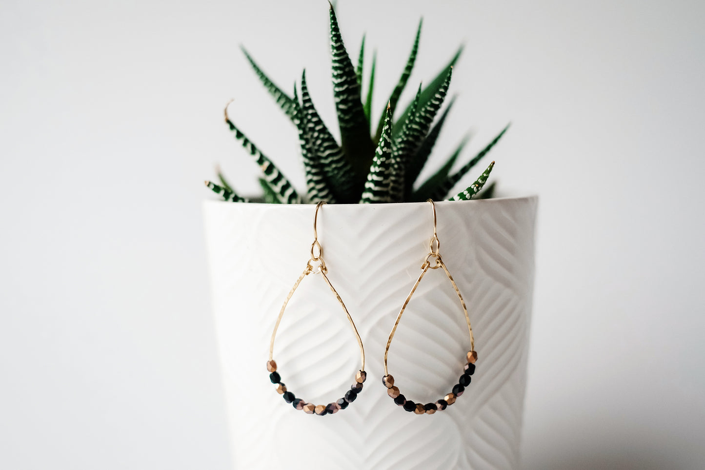 Gold wire teardrop shaped dangle earrings, strung with 12 black and pink speckled faceted glass beads that rest at the bottom of the teardrop shape, hanging from a small white planter containing a green succulent plant.