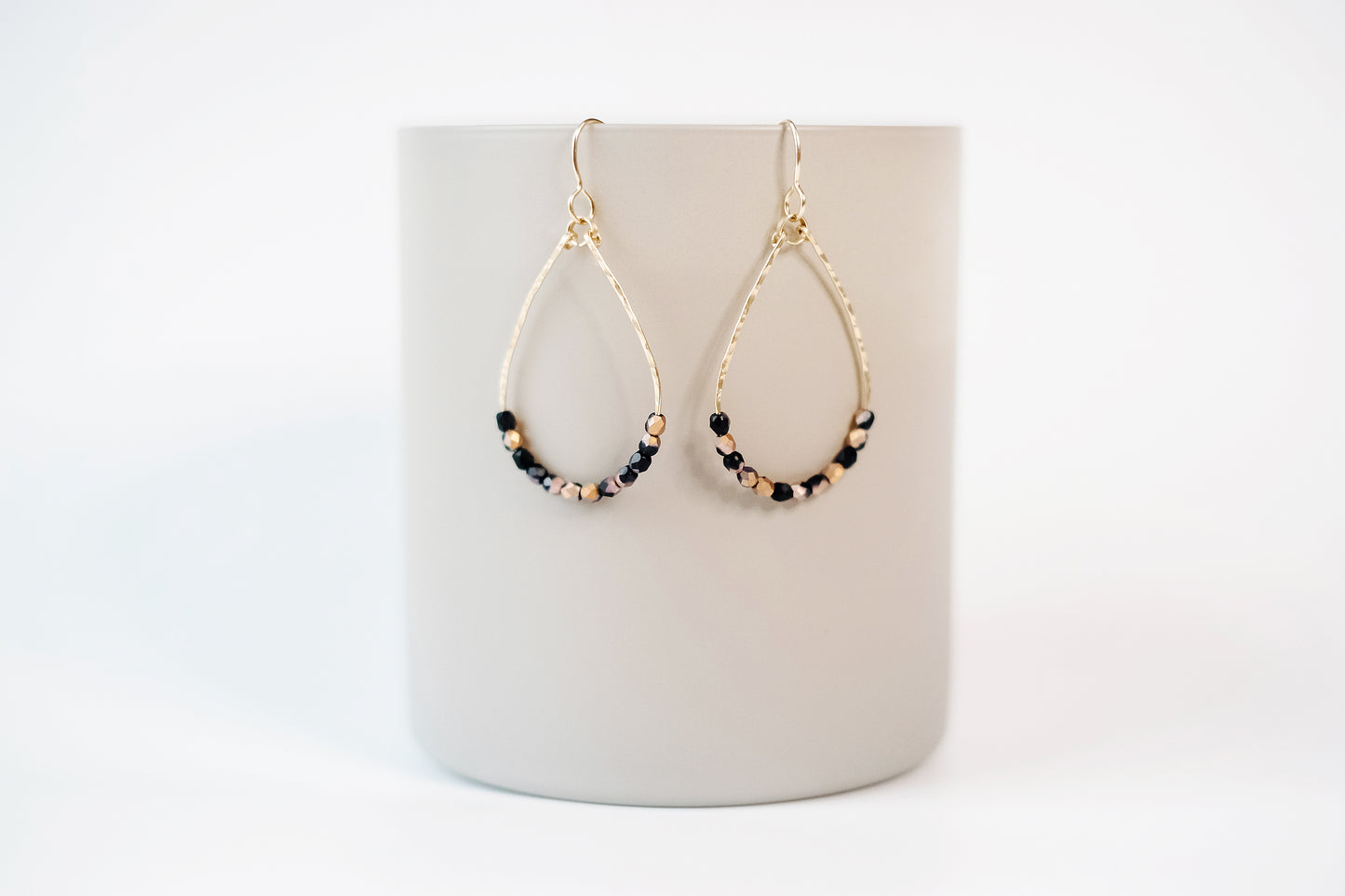 Gold teardrop shaped dangle earrings, strung with 12 black and pink speckled faceted glass beads that rest at the bottom of the teardrop shape, hanging from a beige colored cylinder.