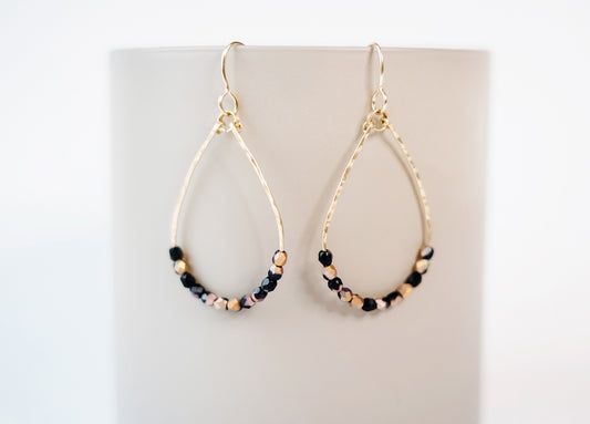 Gold teardrop shaped dangle earrings, strung with 12 black and pink speckled faceted glass beads that rest at the bottom of the teardrop shape, hanging from a beige colored cylinder.