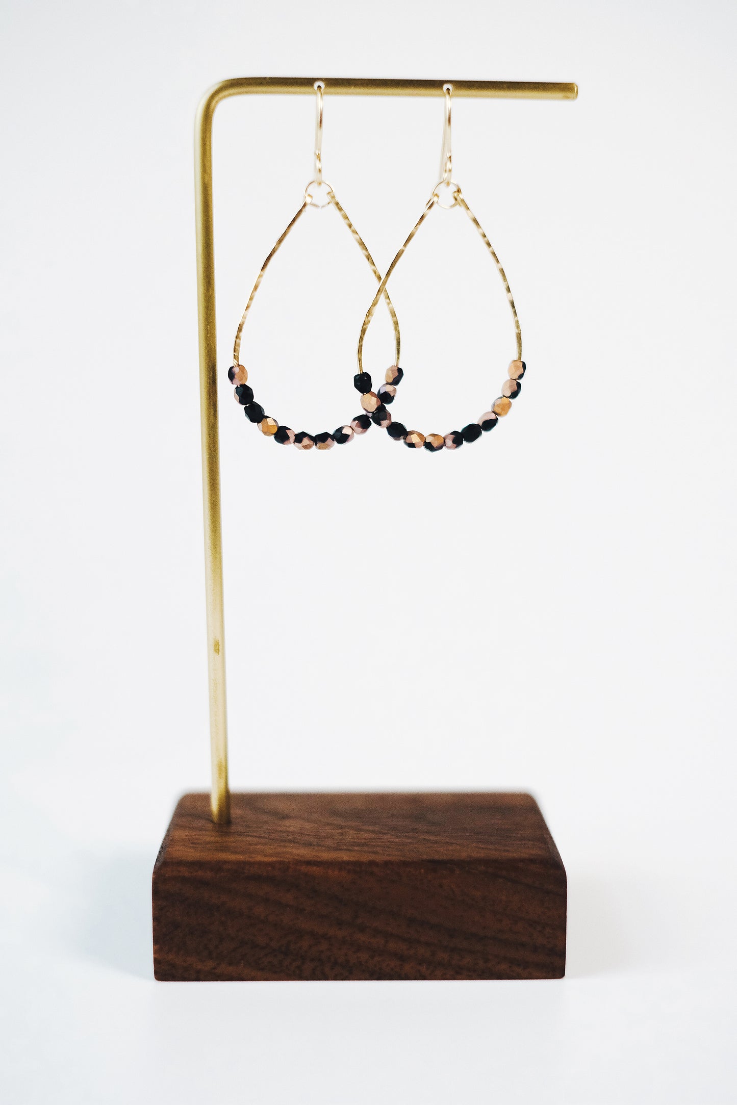 Gold wire teardrop shaped dangle earrings, strung with 12 black and pink speckled faceted glass beads that rest at the bottom of the teardrop shape, hanging from a gold earring hanger with a wooden base..