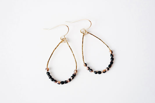 Gold teardrop shaped dangle earrings, strung with 12 black and pink speckled faceted glass beads that rest at the bottom of the teardrop shape, laying flat on a white surface.