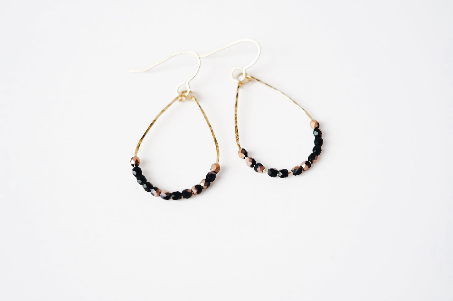 Gold wire teardrop shaped dangle earrings, strung with 12 black and pink speckled faceted glass beads that rest at the bottom of the teardrop shape, laying on a white surface.