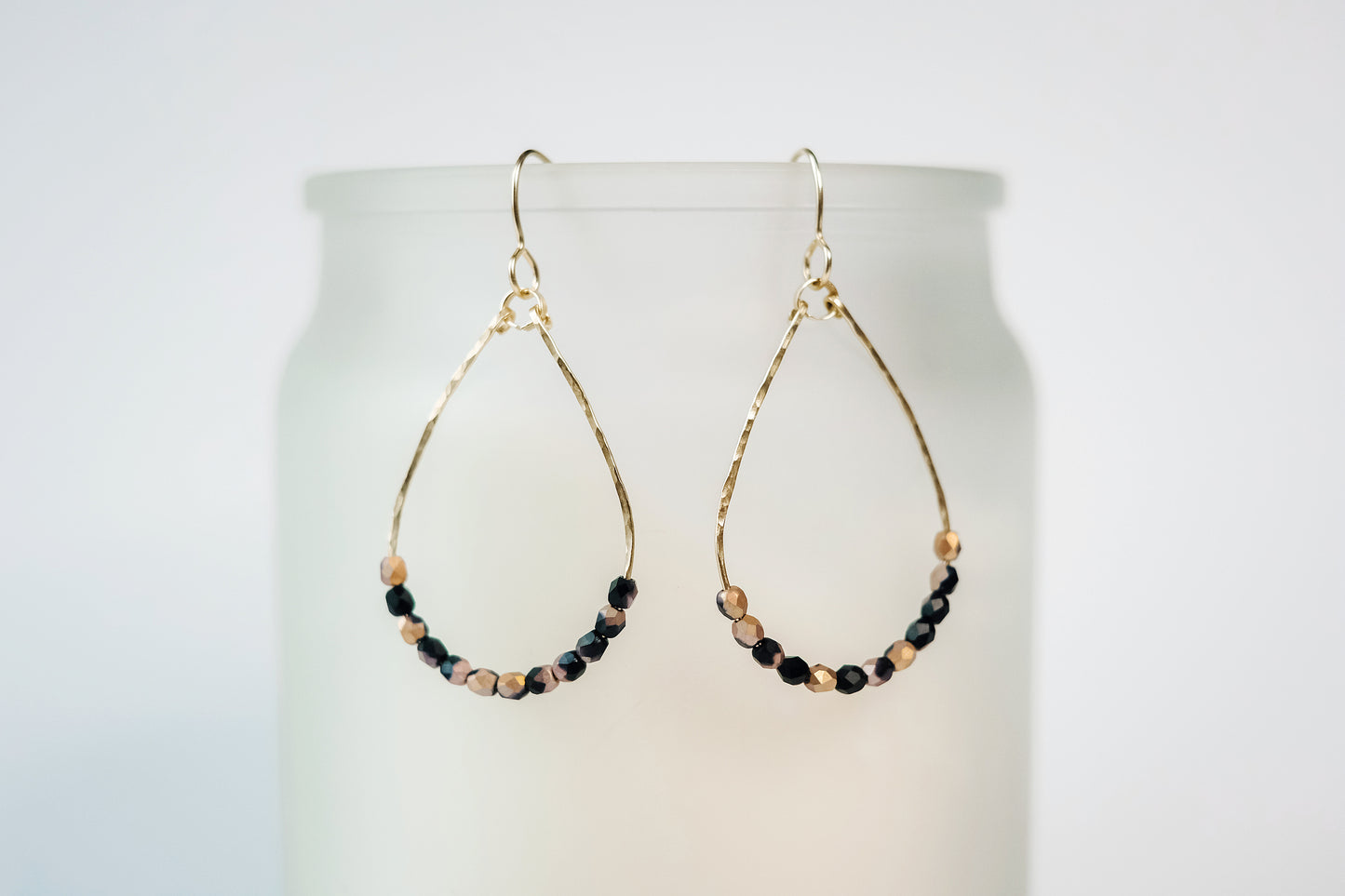 Gold wire teardrop shaped dangle earrings, strung with 12 black and pink speckled faceted glass beads that rest at the bottom of the teardrop shape, hanging from a beige colored cylinder.